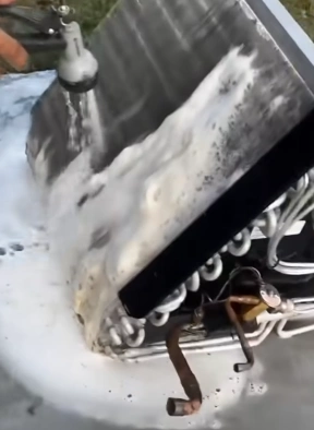 Technician washing and rinsing dirt from AC condenser coil fins during professional cleaning in Bahrain